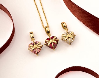 Könnte beinhalten: Drei goldene herzförmige Anhänger mit einer Schleife oben. Die Anhänger sind mit roten, rosa und klaren Kristallen verziert.