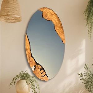 Op de afbeelding: Een ovale spiegel met een natuurlijke houten lijst. Het oppervlak van de spiegel reflecteert een lichtblauwe lucht. De houten lijst heeft een unieke, organische vorm met een lichtbruine kleur. De spiegel hangt aan een crèmekleurige muur.
