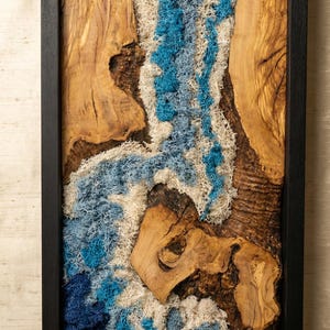 Puede incluir: Arte mural rectangular con marco negro, con base de madera natural. La obra de arte incorpora musgo preservado azul, blanco y azul claro, creando un diseño abstracto texturizado. La veta de la madera añade un elemento natural.