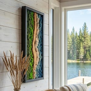 Puede incluir: Una obra de arte mural rectangular con un marco negro. La obra presenta un diseño vertical con texturas verdes, azules y beige, que se asemejan a un paisaje. El arte está montado en una pared de madera blanca, con vistas a un lago y árboles visibles a través de una ventana.
