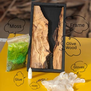 Puede incluir: Una obra de arte rectangular con marco negro que presenta madera de olivo y resina negra. También hay una bolsa de musgo verde, pegamento, ganchos y guantes. La madera de olivo tiene un patrón de vetas natural.