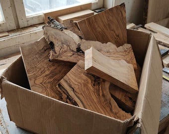 May include: A cardboard box filled with multiple irregular pieces of light brown olive wood. The wood pieces feature prominent dark brown grain patterns, natural edges, and smooth surfaces, showcasing the organic texture of the timber.