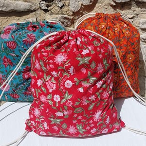 Peut inclure: Trois sacs à cordon de serrage avec des motifs floraux. Les sacs sont rouges, turquoise et orange, chacun avec une fermeture à cordon blanche. Le sac rouge a un motif de fleurs roses et blanches. Le sac turquoise a un motif de fleurs roses et rouges. Le sac orange a un motif de fleurs jaunes et oranges.