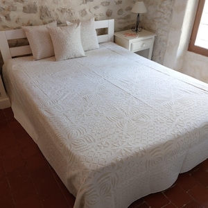 Può includere: Copriletto in cotone bianco con un intricato motivo floreale in rilievo. Il copriletto è su una struttura del letto bianca con tre cuscini bianchi.