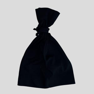 Può includere: Una borsa di stoffa nera, annodata in alto. La borsa è di colore scuro uniforme e sembra essere fatta di un materiale morbido. La borsa è posta su uno sfondo chiaro e uniforme.