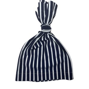 Puede incluir: Un gorro de bebé a rayas blancas y azul marino. El gorro está hecho de tela suave y presenta rayas verticales. La parte superior del gorro está anudada, creando un ajuste elegante y seguro.