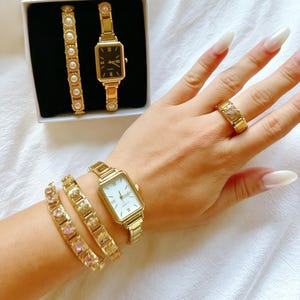 Puede incluir: Un conjunto de relojes dorados que incluye dos relojes rectangulares con esferas blanca y negra, dos pulseras de eslabones con perlas, una pulsera de eslabones con piedras rosas en forma de corazón y un anillo dorado con un corazón rosa.