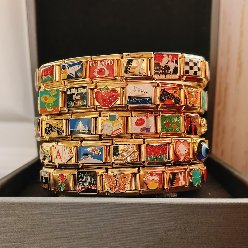Pu&ograve; includere: Un set di braccialetti con ciondoli dorati con ciondoli in smalto colorato. I ciondoli presentano vari disegni, tra cui una tazza di caff&egrave;, un libro, una farfalla, un cupcake e un girasole. I ciondoli sono attaccati a una catena dorata.
