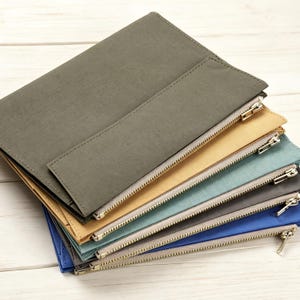 Pode incluir: Uma pilha de cinco bolsas retangulares com zíper nas cores verde-oliva, bege, azul-petróleo, cinza-chumbo e azul-real. Cada bolsa apresenta uma construção plana com um fecho de zíper metálico e uma costura visível no painel frontal.