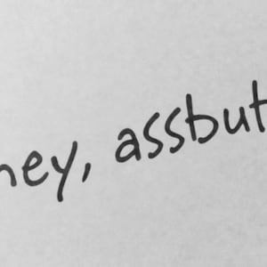 Könnte beinhalten: Eine handschriftliche Nachricht in schwarzer Tinte auf weißem Hintergrund. Der Text lautet "hey, assbutt!" in einer lässigen, leicht ungleichmäßigen Schriftart. Das Ausrufezeichen betont den Satz.