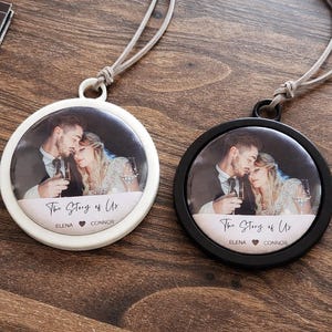 May include: Two circular pendants featuring a couple's photo, one white and one black. Each pendant has a hanging cord and the text "The Story of Us" with names below. They rest on a wooden surface.
