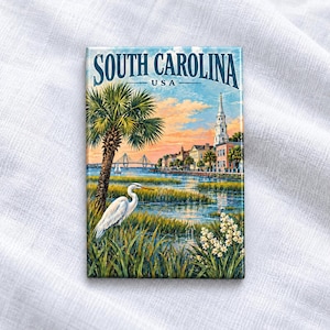 Peut inclure: Un aimant rectangulaire présentant une illustration scénique de la Caroline du Sud, USA. L'image montre une aigrette blanche dans des herbes de marais, un palmier, un pont lointain et un clocher d'église sous un ciel au coucher du soleil.