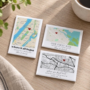 Peut inclure: Trois aimants rectangulaires avec des cartes personnalisées. Un aimant vertical montre une carte de Manhattan avec le texte "where it all began". Deux aimants horizontaux présentent des cartes avec des cœurs rouges, les textes "OUR FIRST KISS" et "where we first met", des coordonnées et des noms.