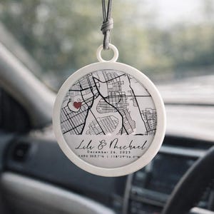 May include: A white, circular car charm with a map design and a red heart. The charm includes the names "Lile & Michael" and the date "December 26, 2025". The charm is hanging from a gray string.