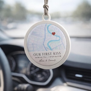 May include: A white, circular car air freshener with a map design and the text "OUR FIRST KISS". A red heart marks a location on the map. Coordinates and names are also printed on the air freshener.
