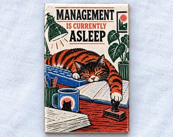 May include: A rectangular magnet featuring an illustration of an orange tabby cat sleeping on a blue computer keyboard and an open notebook. The text reads MANAGEMENT IS CURRENTLY ASLEEP. Includes a desk lamp, a mug, pens, and a rubber stamp.