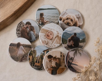 May include: A collection of nine circular photo magnets featuring various printed images, including a child at the beach, a dog, a couple, a baby, a family, a person in a field, a building, and a sunset beach scene, arranged in a cluster.