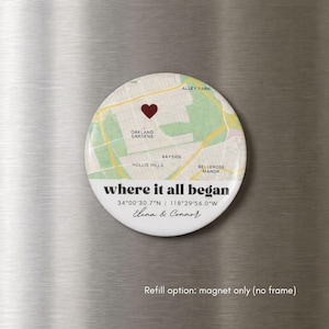 May include: A circular magnet with a map design, a red heart, and the text "where it all began." The magnet includes coordinates and names, with a white border. The background is a metallic surface.