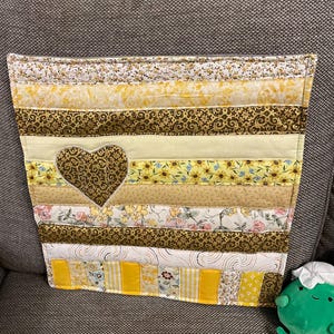 May include: A patchwork wall hanging with a heart-shaped cut-out. The design incorporates strips of fabric in shades of yellow, brown, and floral patterns. The heart is a solid brown fabric. The item is displayed on a grey sofa.