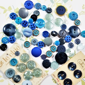 May include: Assortment of blue, gray, and white buttons in various sizes and styles. Some buttons have a textured surface, while others are smooth. The buttons are arranged in a random pattern on a white surface. The text "29c le Chic" and "Guarant Wash" is visible on a white card.