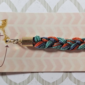 May include: A braided bracelet with orange, teal, and navy blue threads. The bracelet has a gold-toned clasp and a tag that reads "Bracelet Bar" and "Clip me to your favorite stackable clasp!"