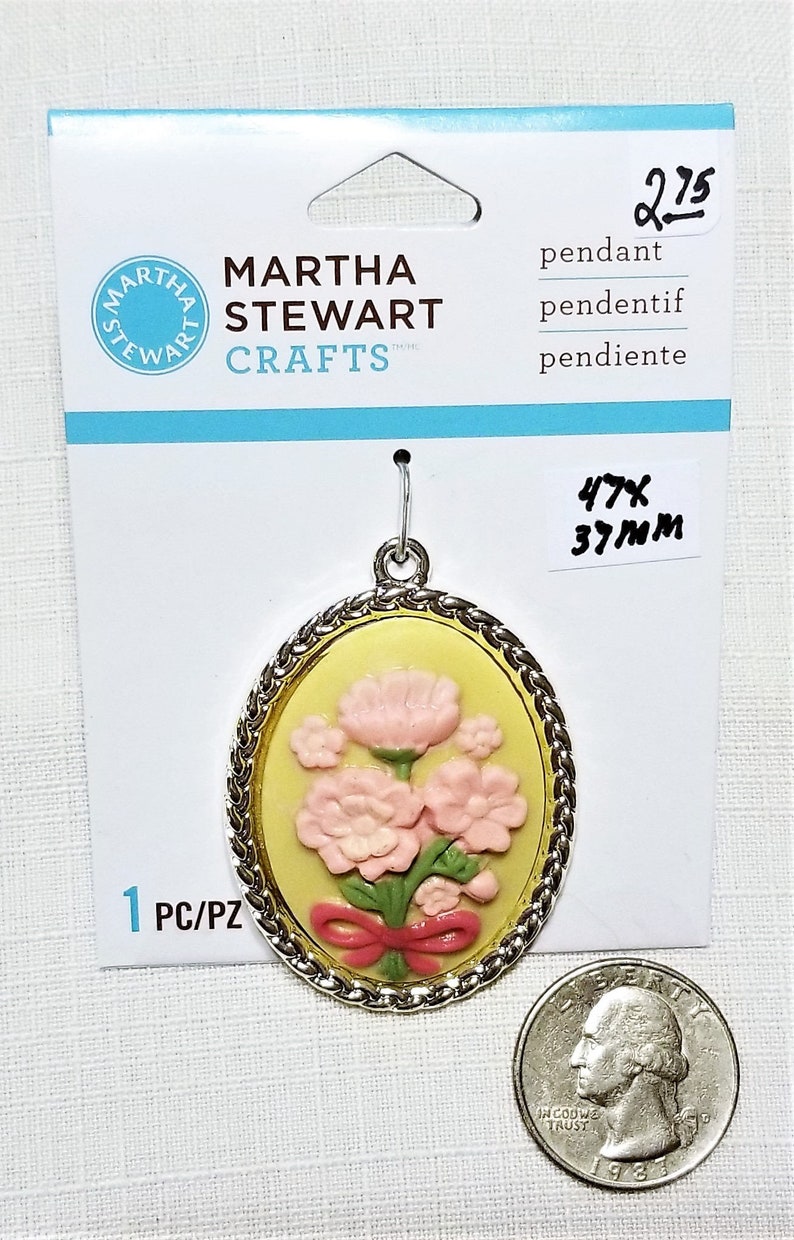 May include: A silver-toned pendant with a yellow oval cameo featuring a pink flower bouquet with a red bow. The pendant is 1.85 inches (4.7 cm) tall and 1.46 inches (3.7 cm) wide. The packaging includes the text "MARTHA STEWART CRAFTS" and "pendant, pendentif, pendiente".