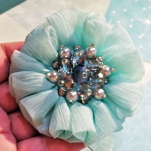 May include: A light blue fabric flower with a center made of iridescent blue and clear rhinestones and pearl-like beads.