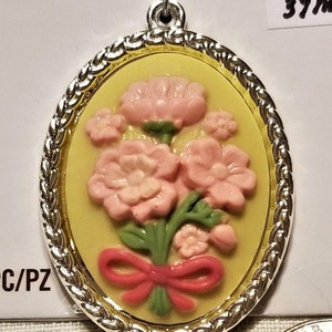 May include: A yellow oval pendant with a silver rope border and a gold inner border. The pendant features a pink flower bouquet with a red bow.