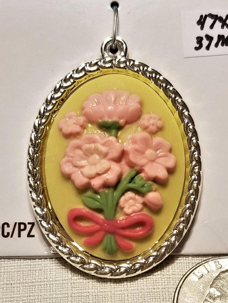May include: A yellow oval pendant with a silver rope border and a gold inner border. The pendant features a pink flower bouquet with a red bow.