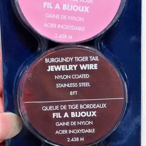 May include: Three spools of nylon coated jewelry wire in pink, brown, and silver. Each spool is labeled with the color, "Jewelry Wire", "Nylon Coated", "Stainless Steel", "8 ft", "Queue de Tige", "Fil a Bijoux", "Gaine de Nylon", "Acier Inoxydable", and the measurement in feet and meters.