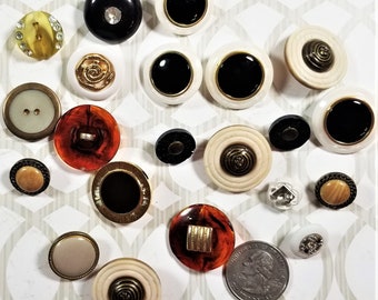 May include: Assortment of vintage buttons in various colors, patterns, and materials, including white, black, brown, gold, and ivory. Some buttons have a raised design, while others are flat. The buttons are arranged on a white surface with a patterned background.