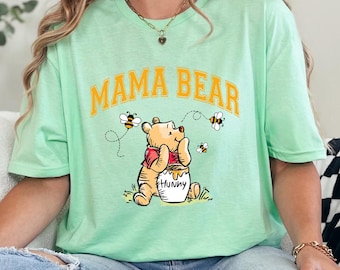 May include: A light green short-sleeve t-shirt featuring the text MAMA BEAR in yellow arched letters. Below the text is a graphic of a yellow bear wearing a red shirt, holding a white honey pot labeled HUNNY, surrounded by small bees.