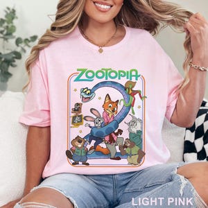 Pode incluir: T-shirt rosa claro com a palavra "ZOOTOPIA" em verde e um desenho animado de personagens do filme. O desenho animado inclui uma cobra, uma raposa, um coelho e outros animais. As palavras "LIGHT PINK" est&atilde;o na parte inferior.