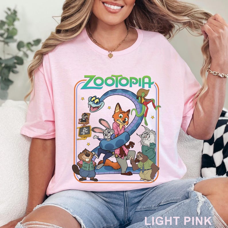 Pode incluir: T-shirt rosa claro com a palavra "ZOOTOPIA" em verde e um desenho animado de personagens do filme. O desenho animado inclui uma cobra, uma raposa, um coelho e outros animais. As palavras "LIGHT PINK" est&atilde;o na parte inferior.