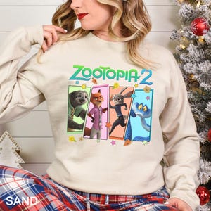May include: A sand-coloured sweatshirt with the text "ZOOTOPIA 2" in green and blue. The design features four rectangular panels with cartoon characters. The sweatshirt has long sleeves and a crew neck.