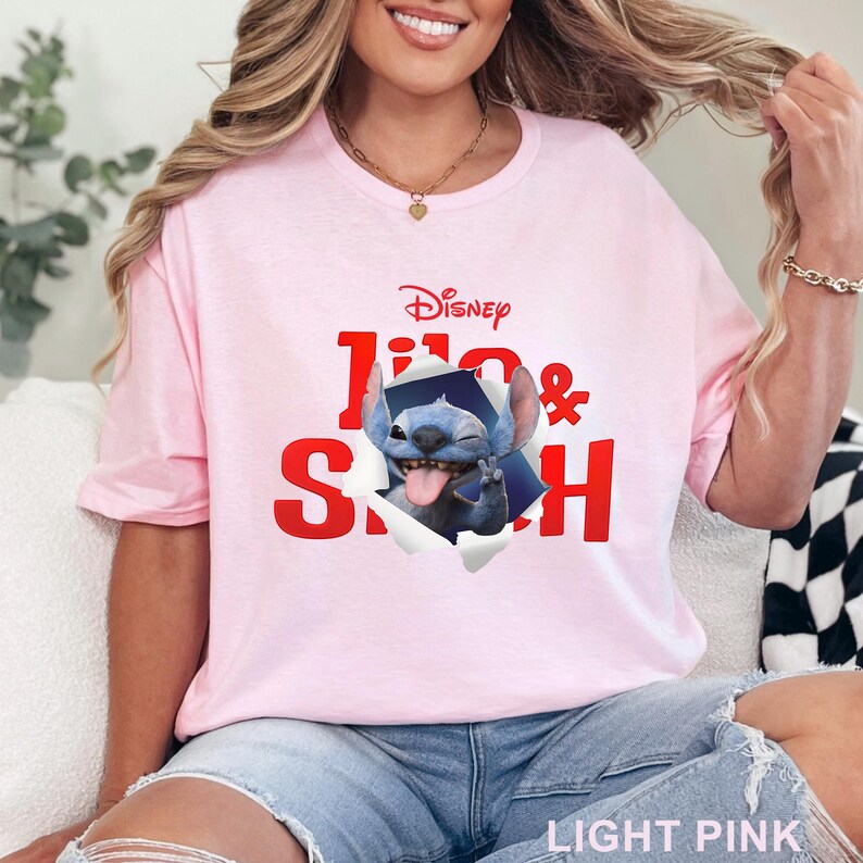Puede incluir: Camiseta rosa claro con el logotipo de Disney y las palabras "Lilo & Stitch" en rojo. El gr&aacute;fico muestra a Stitch saliendo de un efecto de papel rasgado. La camiseta est&aacute; hecha de un material suave.