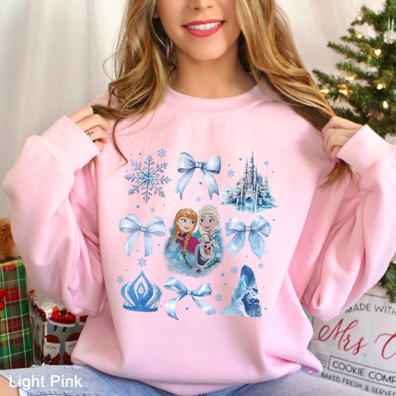 Puede incluir: Sudadera rosa claro con un dise&ntilde;o de copos de nieve, lazos e im&aacute;genes de personajes de una pel&iacute;cula de animaci&oacute;n popular. El dise&ntilde;o incluye un castillo, una corona y el texto "Light Pink" en la parte inferior.