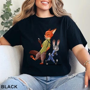 Pode incluir: Camiseta preta com uma ilustra&ccedil;&atilde;o de desenho animado de Nick Wilde e Judy Hopps de Zootopia. Nick, uma raposa, est&aacute; segurando um telefone e Judy, uma coelha, est&aacute; na coleira. A palavra "BLACK" est&aacute; impressa na parte inferior.