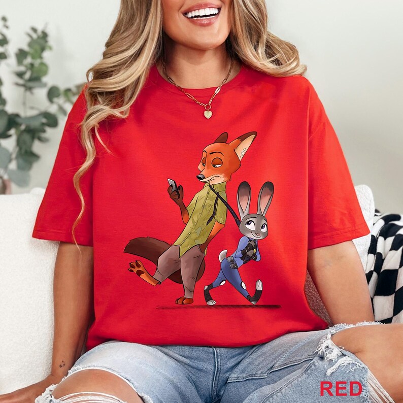 Pode incluir: Uma camiseta vermelha com uma ilustra&ccedil;&atilde;o de desenho animado de Nick Wilde e Judy Hopps de Zootopia. Nick, uma raposa, est&aacute; segurando um telefone, enquanto Judy, uma coelha, est&aacute; em um uniforme de policial. A palavra "RED" &eacute; impressa no canto inferior direito.