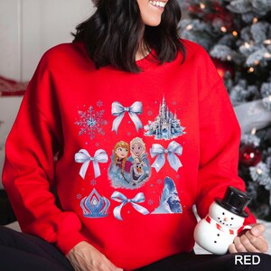 Puede incluir: Una sudadera roja con un dise&ntilde;o de tem&aacute;tica invernal. El dise&ntilde;o incluye im&aacute;genes de un copo de nieve, lazos, un castillo y personajes. La palabra "RED" est&aacute; impresa en blanco en la esquina inferior derecha de la sudadera.