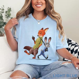 Pode incluir: Camiseta azul clara com uma ilustra&ccedil;&atilde;o de desenho animado de Nick Wilde e Judy Hopps de Zootopia. Nick, uma raposa laranja, usa uma camisa verde, e Judy, uma coelha cinza, est&aacute; com um uniforme azul. O texto "LIGHT BLUE" est&aacute; na parte inferior.