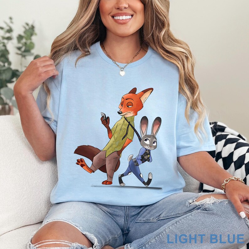 Pode incluir: Camiseta azul clara com uma ilustra&ccedil;&atilde;o de desenho animado de Nick Wilde e Judy Hopps de Zootopia. Nick, uma raposa laranja, usa uma camisa verde, e Judy, uma coelha cinza, est&aacute; com um uniforme azul. O texto "LIGHT BLUE" est&aacute; na parte inferior.