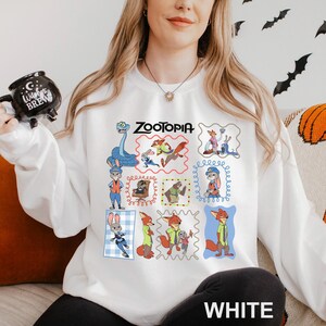 May include: White sweatshirt featuring a colorful collage of Zootopia characters. The word "Zootopia" is printed in black above the images. The word "WHITE" is printed in white at the bottom of the sweatshirt.