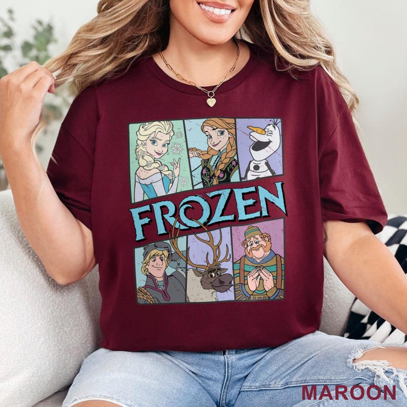 May include: Maroon t-shirt featuring a graphic of characters from the movie "Frozen". The design includes Elsa, Anna, Olaf, Kristoff, Sven, and a character in a green outfit. The word "FROZEN" is in blue letters.