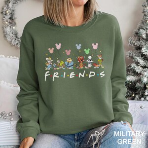 May include: An olive green sweatshirt featuring a cartoon design of various characters holding Mickey Mouse-shaped balloons. The word "FRIENDS" is printed in white with colorful dots. The bottom right corner of the sweatshirt has the text "MILITARY GREEN".
