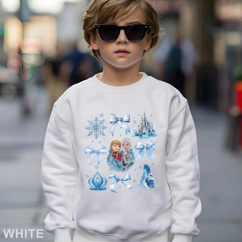 Puede incluir: Sudadera blanca con un gr&aacute;fico colorido de los personajes Anna y Elsa de la pel&iacute;cula Frozen, junto con copos de nieve, lazos y un castillo. Adecuada para ni&ntilde;os.
