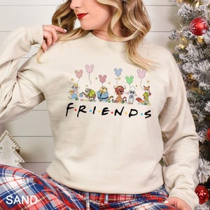 May include: Sand-colored sweatshirt featuring the word "FRIENDS" in a colorful font, with cartoon characters holding Mickey Mouse-shaped balloons. The sweatshirt has long sleeves and a crew neck.