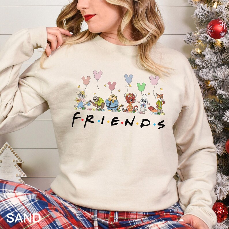 May include: Sand-colored sweatshirt featuring the word "FRIENDS" in a colorful font, with cartoon characters holding Mickey Mouse-shaped balloons. The sweatshirt has long sleeves and a crew neck.
