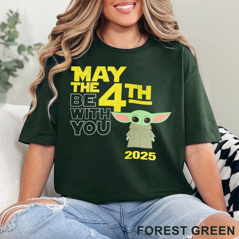 May include: A dark green t-shirt with the text "May the 4th Be With You 2025" in yellow. A cartoon image of a green alien character is in the centre of the shirt.