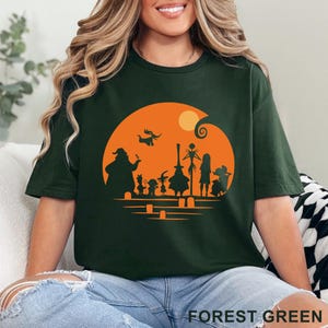 Pu&ograve; includere: T-shirt verde foresta con un design a silhouette a tema Halloween. Il design presenta personaggi ed elementi su un cerchio arancione con la luna piena. Il testo "FOREST GREEN" &egrave; stampato in basso sulla maglietta.